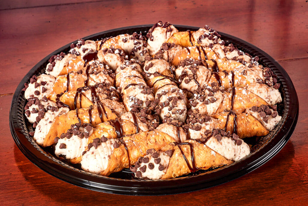A catering tray of chocolate chip cannoli covered in powdered sugar and chocolate sauce