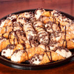 A catering tray of chocolate chip cannoli covered in powdered sugar and chocolate sauce