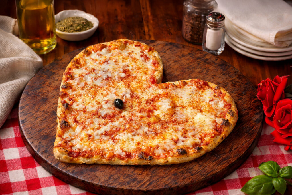 A heart-shaped cheese pizza served on a rustic wooden board with red roses and a checkered tablecloth.
