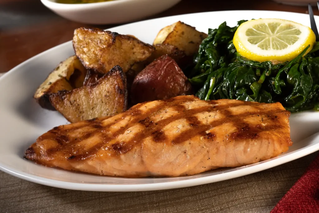 Grilled salmon fillet with char marks, served with roasted potatoes, sautéed spinach, and a fresh lemon slice on a white plate.