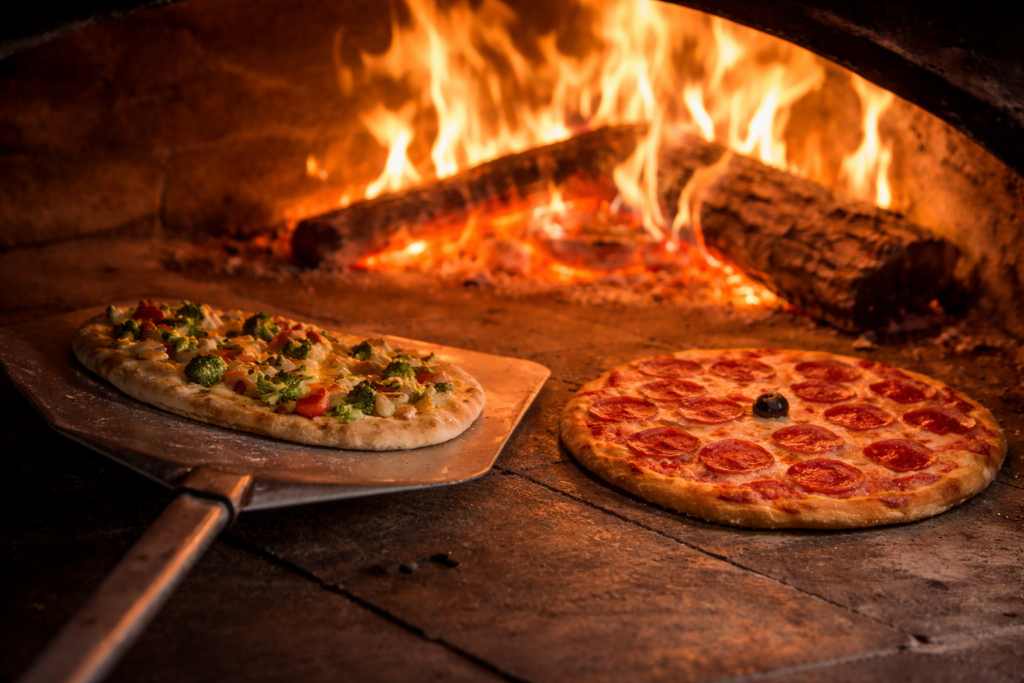 Two pizzas baking inside a rustic wood-fired brick oven, flames glowing in the background. A vegetable pizza sits on a metal peel to the left, while a pepperoni pizza with a single black olive in the center rests directly on the stone oven floor.