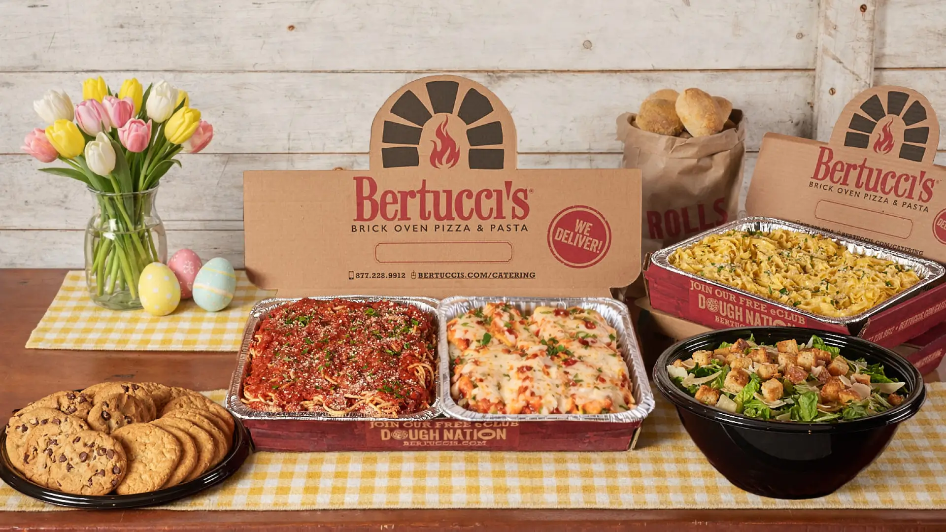 Bertucci’s catering spread with trays of baked pasta, spaghetti with marinara, Caesar salad, rolls, and chocolate chip cookies on a table with spring flowers and Easter eggs.