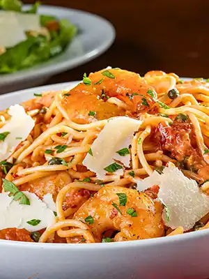 Shrimp Rossini with sautéed shrimp, spaghetti, tomatoes, capers, fresh herbs, and shaved Parmesan in a white bowl.
